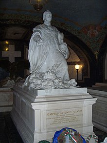 White marble tomb in a dark crypt depicting a sitting man with a moustache, his hat in one hand, his other hand extended over the Holy Crown of Hungary.