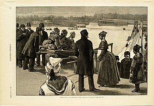 A winter scene. Men, women, and children are at the top of a hill, boarding toboggans preparing to slide down one of two tracks. Horses pulling sleds are visible in the distance, along with several buildings.
