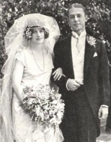 A young white bride and groom, standing arm in arm and smiling; dark-haired bride has a voluminous veil and a white gown with short sleeves; she is holding a bouquet; groom is in formal dress and wearing a corsage; his hair is side-parted and short