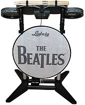 A drum set video game controller with four elevated drum pads mounted on a frame, along with a bass pedal attached to a bottom crossbar. A thin panel, stating "Ludwig" and "The Beatles" is mounted to the front. The drum pads are colored in a metallic gray pattern, while most of the rest is either black or gray.