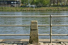 An obelisk of weathered stone, about two feet high and marked with the letters "UBR" on a relief, stands before a three-pipe fence separating the viewer from the River Thames.