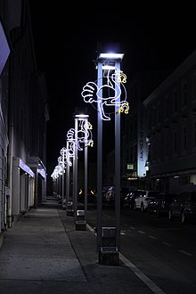 Eine Vielzahl an Straßenlaternen entlang einer asphaltierten Straße bei Nacht. An jeder Laterne hängt ein in LEDs konturierter Vogelstrauß mit Hufeisen.
