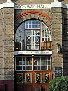 The main entrance with a narrow arch rising to the top of the upper floor. The arch is flanked by two columns in silver-grey brick while the rest of the building is varied with red-brick detailing. There is a lot of glass in the façade.