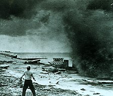 A man stands on a crumbled roadway by a body of water. The road is filled with bricks and pieces of broken concrete lying around. The man is tense, facing a large, black, smoke-like wave of water that fills the upper right-hand portion of the picture. The man looks frightened and his legs are bent, spaced apart, as if preparing to run or sprint.