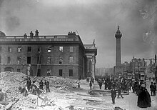 "Black and white photograph of a Dublin street showing a ruined building to the left with rubble to the foreground and pedestrians milling around the main street below a large pillar with statue on top"