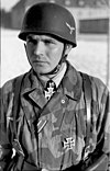 A man wearing a camouflage military uniform, steel helmet and a neck order in the shape of a cross.