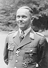 A man wearing a military uniform and neck order, in the shape of a cross. He has short hair that is combed back and a determined facial expression.