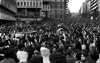 A large crowd, many people with fists raised, watches a hearse passing