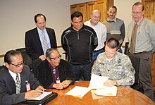 Three men sitting and four men standing while one of them signs a document.