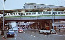 A white-and-green monorail train hangs over a city street with several cars.
