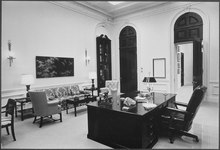 A black and white photo of a tall white room with detailed moldings and tall arched doors. A large desk sits just off center with a chair behind it and a small seating area with a couch in front of it.