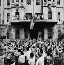 A photograph of celebratory crowds in front of and on a building draped by an American flag at the liberation of a POW camp.