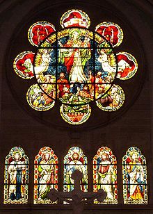 A rose window of simple form having a circle with eight lobes like a flower. It has glass showing Christ rising into heaven watched by apostles and angels. Below are five arched windows each with a saint. Red and yellow are the predominant colours