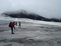 Three climbers descending a glacier on a cloudy day with a rocky mountain peak in the background.