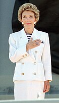Portrait of a woman standing with her hand on her heart, wearing a white 1980s style suit with large shoulder pads and gold buttons.