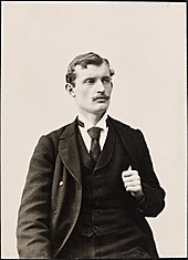 Frontale Schwarzweißfotografie von einem Mann, der nach rechts an der Kamera vorbeiblickt. Er trägt einen Schnurrbart und einen Anzug, in den er seinen linken Daumen steckt.