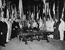 Franklin D. Roosevelt and three other men seated at a table, surrounded by many other men and flags