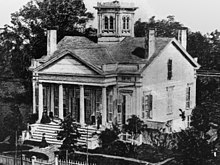 A white house with multiple chimneys and a tower. People are on the front porch and someone is arriving at the home and about to embark up the staircase. The front porch has a roof over a set of four columns. Trees are visible on the lawn.