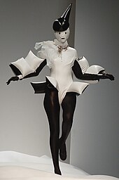 A model walks on a runaway wearing a white dress with black stalkings. The dress has inflatable small white cushions on it, and the model is wearing a white and black mask.