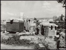 Monochromatic image showing four male persons inspecting two massive sarcophagi in the midst of ancient ruins. A town, a low church steeple, and a hill appear in the background.