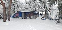 A structure made of plywood, tarps and corrugated metal in the snow with a homeless man working next to it.