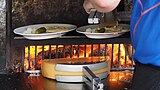 A half raclette cheese being melted on a wood fire while the other half is being scraped from its melted part in a plate.