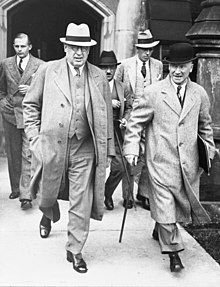 Two men in the foreground walk down a sidewalk, followed by three men in the background. The man in the left of the foreground is portly and wears a fedora, round-rimmed glasses, and a three piece suit. The man on the right is wearing an overcoat and a bowler hat, and holds a cane in his right hand and some documents in his left.