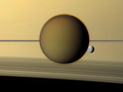 An image of Titan in front Saturn as the smaller moon Dione passes behind its fellow moon. The north pole of Titan is covered by an atmospheric hood which appears as a detached layer at the top of the moon. Imaged on May 21, 2011, by the Cassini spacecraft.[226]