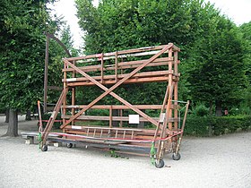Seitliche Farbfotografie eines zweistöckigen Holzgestells mit Schutzgeländern auf Rädern. Links ist eine Konstruktion als Viertelkreis angebracht. Im Hintergrund sind ein Weg und Bäume.