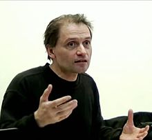 The picture depicts the top half of Michael Heinrich who is wearing a black long-sleeve shirt. His hands in front of him and he is giving a lecture.