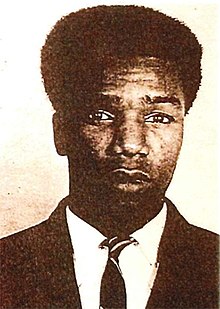 A sepia-toned image of an African-American man with a slight Afro, wearing a jacket and dark necktie, looking at the camera without smiling