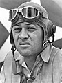 Head of a squinting man wearing a shirt unbottoned at the collar and a cloth aviator's cap with headphones built into the ear flaps, an unbuckled chin strap, and goggles pushed up onto his forehead.
