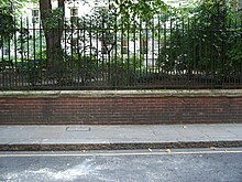 A brick wall topped by metal railings stands at the side of a tarmac road. Behind the wall, grass and trees are visible, growing at the height of the top of the wall.