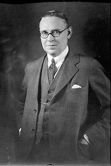 Studio portrait of Richard Gavin Reid. He has an oval, fleshy face with receding hair combed back, a short nose, small shaply mouth. His expression is quizzical and he is compressing a smile. He wears round horn-rimmed glasses and has his hands in the jacket pockets of his neat three-piece suit.