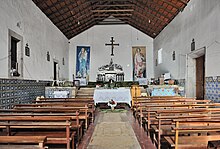 Igreja de Nossa Senhora do Rosário. Schlichter, einschiffiger Kirchenraum mit hölzernen Kirchenbänken links und rechts und zwei Wandbildern links und rechts eines schlichten Kreuzes.
