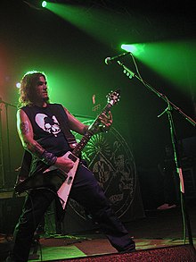 A man in a black sleeveless shirt plays the guitar on a stage.