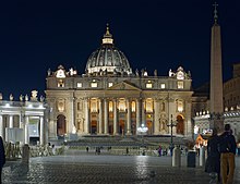 Frontale Nachtfotografie vom Petersdom mit wenigen Menschen auf dem Platz im Vordergrund. Der Dom mit seinem Säuleneingang und der Kuppel ist beleuchtet. An der rechten Seite ist ein dunkler Obelisk mit einer Kreuzspitze zu erkennen.
