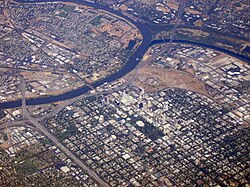 High-angled aerial shot of a developed city, a suburban grid dominates the lower half of the image. A river bisects the city from the left before forking; the first fork continues up and to the right edge of the image; the second curves up and around to finish on the left, enclosing industrial units and other domestic properties.