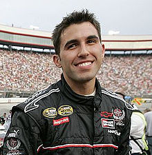 Almirola, with spiked hair, smiles at the camera while wearing his black racing suit. The filled grandstands are in the background.