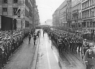 Soldiers with rifles and helmets march along a road past a reviewing party and guard of honor
