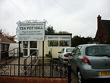 A flat-roofed temporary building, neatly finished in cream. A ramp for 'disabled access' leads to the door, and two cars are parked on the right. Prominent on the left a sign decorated with 'wrought' iron scrolls reads Bottesford town council, TEA POT HALL, pop-in room for the benefit of senior citizens