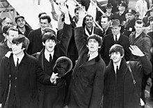 The Beatles are standing in front of a crowd of people at the bottom of an aeroplane staircase.