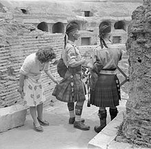 Two Highland-regiment pipe majors in kilts, Glengarry bonnets, and undress army blouses with insignia, as well as leather shoes and hose with flashes; a woman in a white skirt is examining the pleats of one of the kilts.