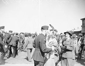 Man in dark military uniform in the arms of a female civilian, among a crowd of other civilian and military people
