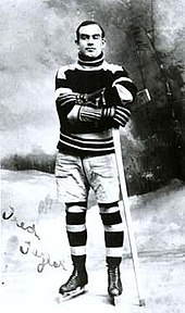 A young man poses for a photo, wearing a sweater, skates, and holding an ice hockey stick on his left side