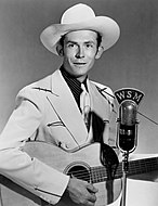 A photograph of a young man wearing a cowboy hat, holding a guitar and standing next to a microphone