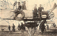 An airship gondola is shown in close-up from mid-left to mid-right; several items of equipment hang from the gondola's side; below the gondola a pyramid of steel tubing protrudes, shielding a cylindrical fuel tank; four crew members are in the gondola; above it mooring ropes and a rip-line are visible. A flat-ended propeller is also visible on the right