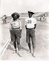 1904 marathon participants Tau and Mashiani stand in a stadium looking at the camera, both wearing hats