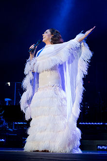 A woman in a white dress with a white stole, with her right hand stretched and a microphone in her left hand, looking to the side.