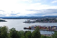 Farbfotografie in der Obersicht von einem Fjord mit Hafen. Die Wasserfläche erstreckt sich vom vorderen Bereich bis zum linken Hintergrund. Der vorderste Pier besteht aus Containern und die hinteren Piers liegen neben den ersten Häusern der Stadt.